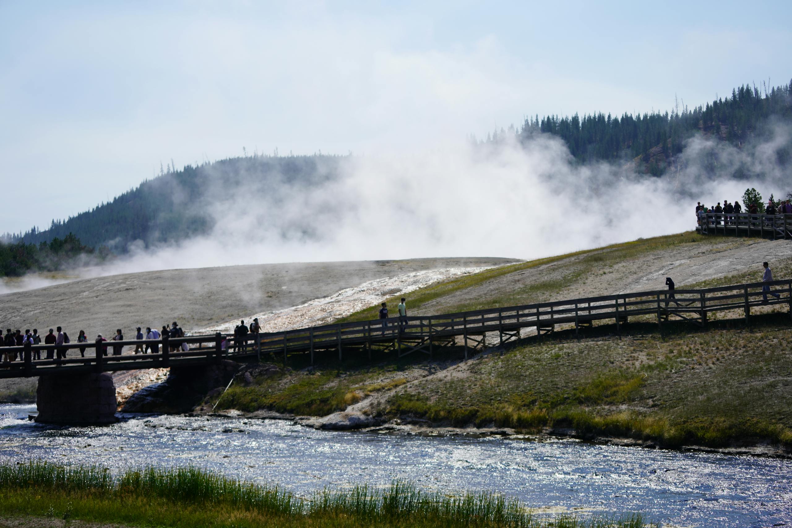 Tourists exploring a footbridge amidst steam in Yellowstone's geothermal landscape.